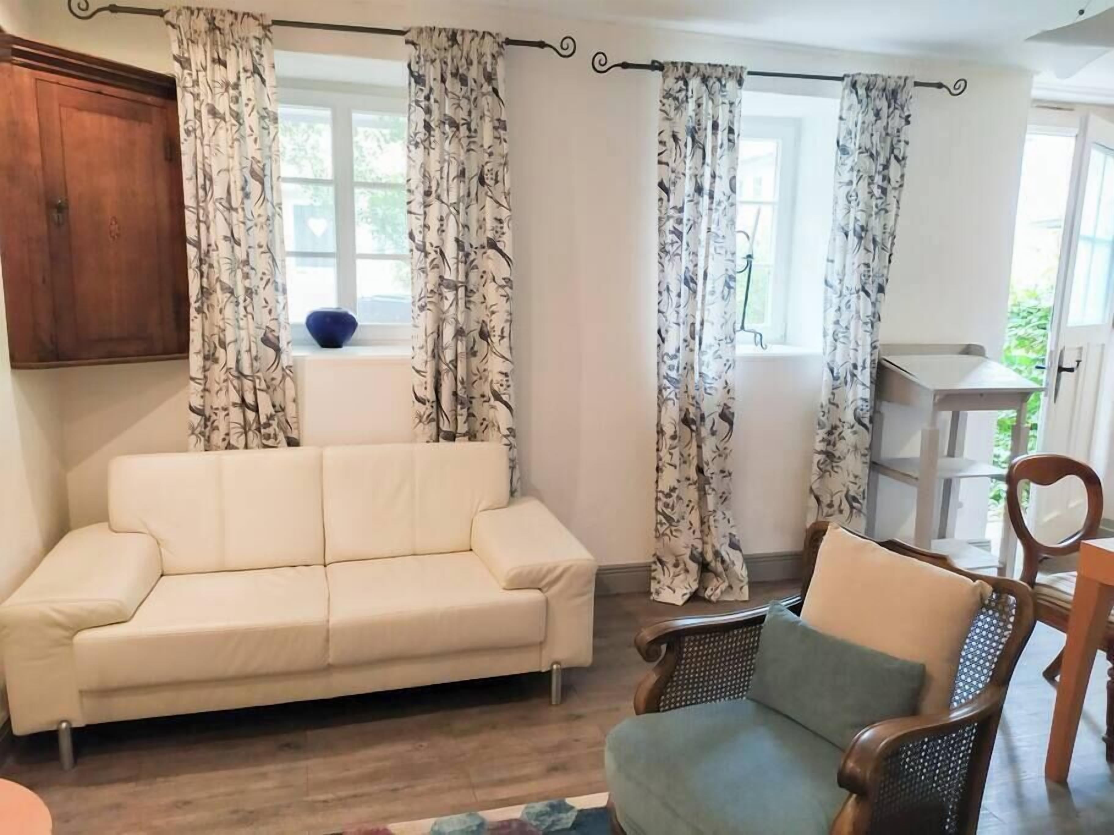 Ein Wohnzimmer mit einer Couch und einem Sessel, einem Holzschrank und einem Fenster mit Vorhängen.