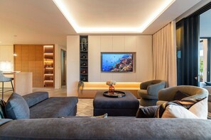 A modern living room with a flat-screen TV, grey sofas, a coffee table, and a kitchen area with wooden cabinets and a built-in shelf. - Walk to beach, Pool, Camps Bay -Terrace Royale Apt (Cape Town)