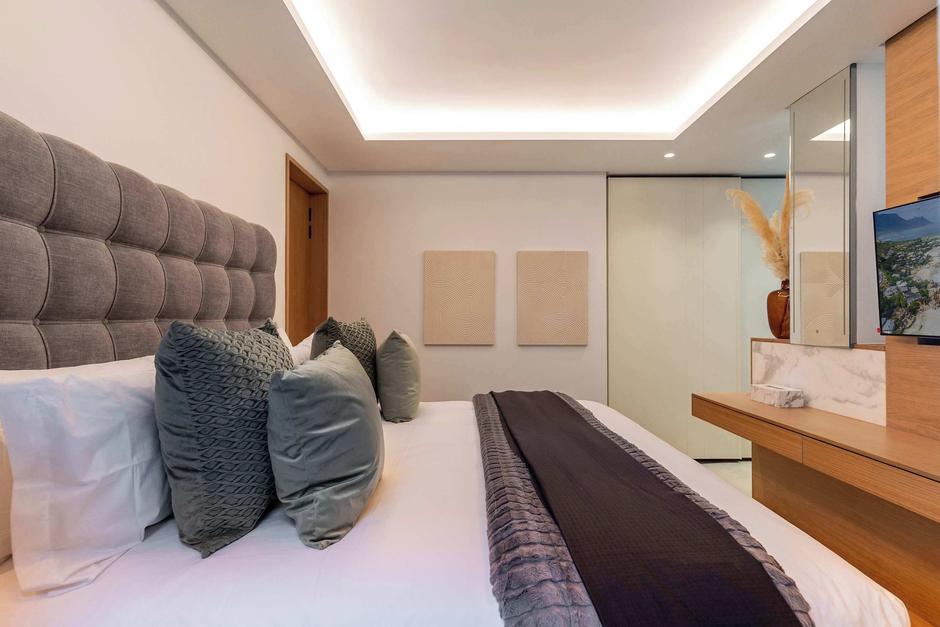 Une chambre moderne avec une tête de lit capitonnée, un lit agrémenté de plusieurs coussins, une table de chevet, un miroir et des œuvres d’art murales.