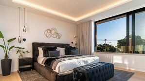 Quarto moderno com uma cama grande, mesas de cabeceira, uma planta em vaso e vista para o exterior.