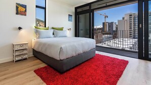 A modern bedroom with a large bed, a red rug, and a balcony with a city view. - City Views, Shared Gym, Cape Town -York Cityscape Apt (Cape Town)