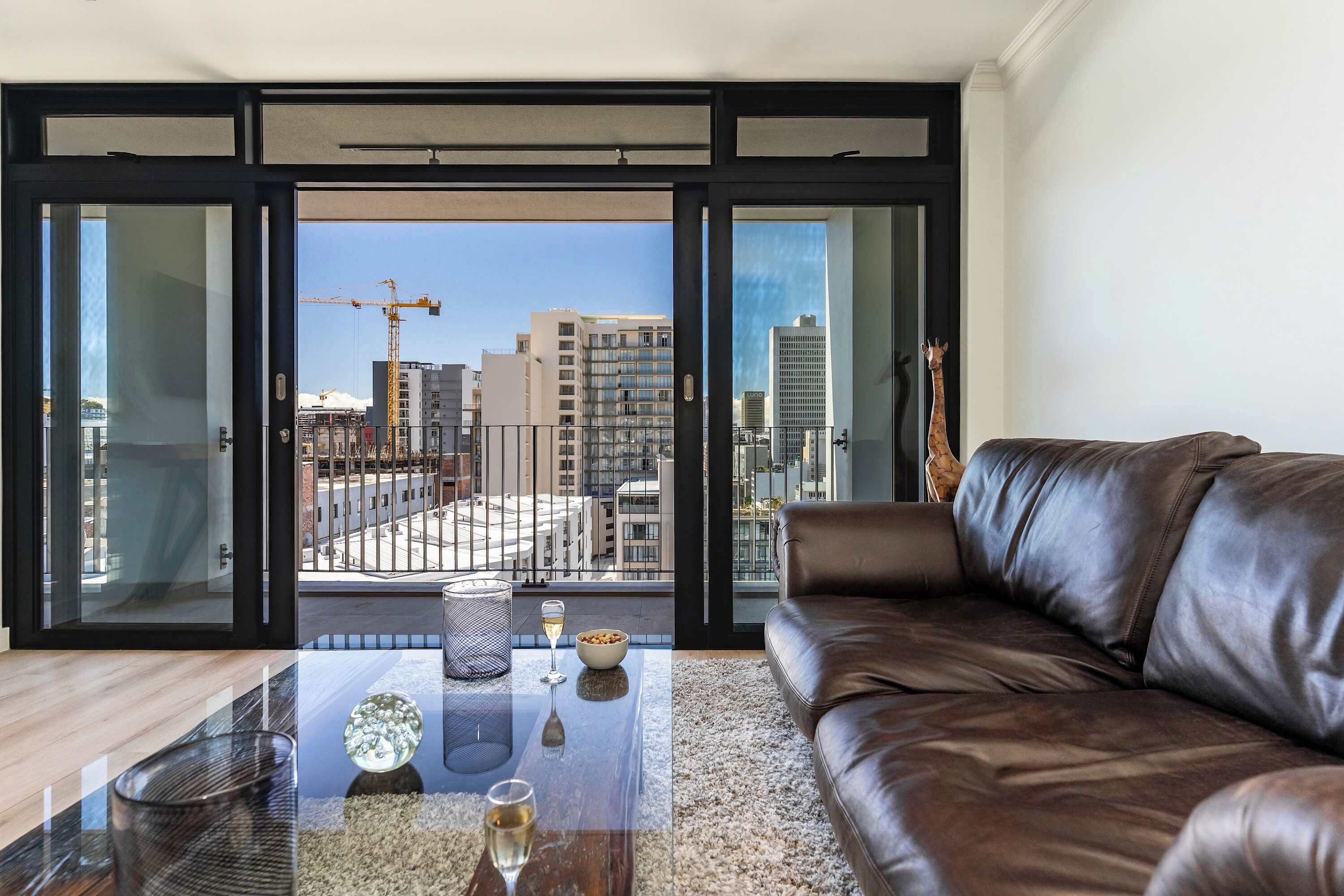 Ein modernes Wohnzimmer mit Glastür zum Balkon, einem Ledersofa, einem Couchtisch mit Dekoration und Blick auf die Stadtlandschaft.
