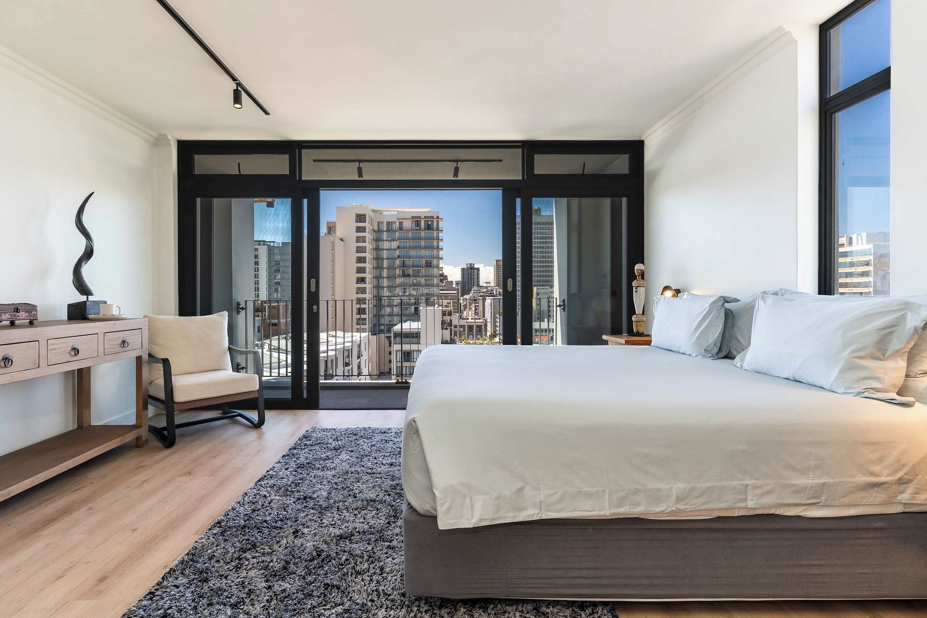 A modern bedroom with a large bed, a desk, and a chair. It has a city view through a glass door.