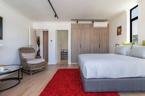 Ein modernes Schlafzimmer mit einem großen Bett, einem roten Teppich, einem Korbstuhl und Blick auf die Stadt durch ein Fenster.