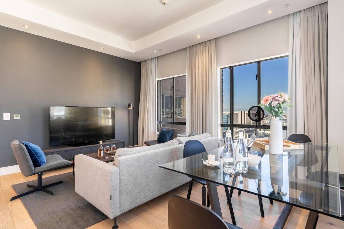 Een moderne woonkamer met een flatscreen televisie, grijze banken, een glazen eettafel en grote ramen met uitzicht op de stad.