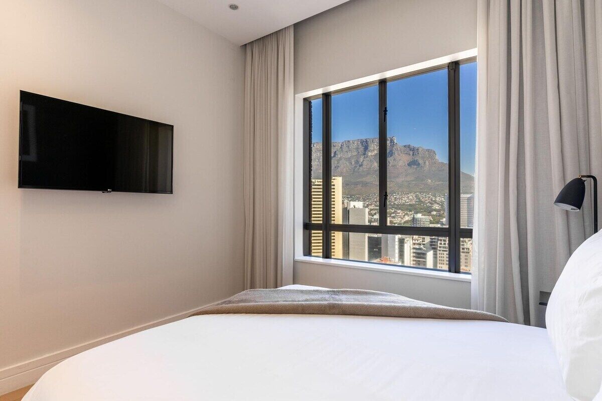 Een hotelkamer met een bed, een aan de muur bevestigde televisie, een groot raam met gordijnen en uitzicht op een stadsbeeld en bergen.