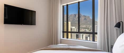 A hotel room with a bed, a TV mounted on the wall, a large window with curtains, and a view of a cityscape and mountains.