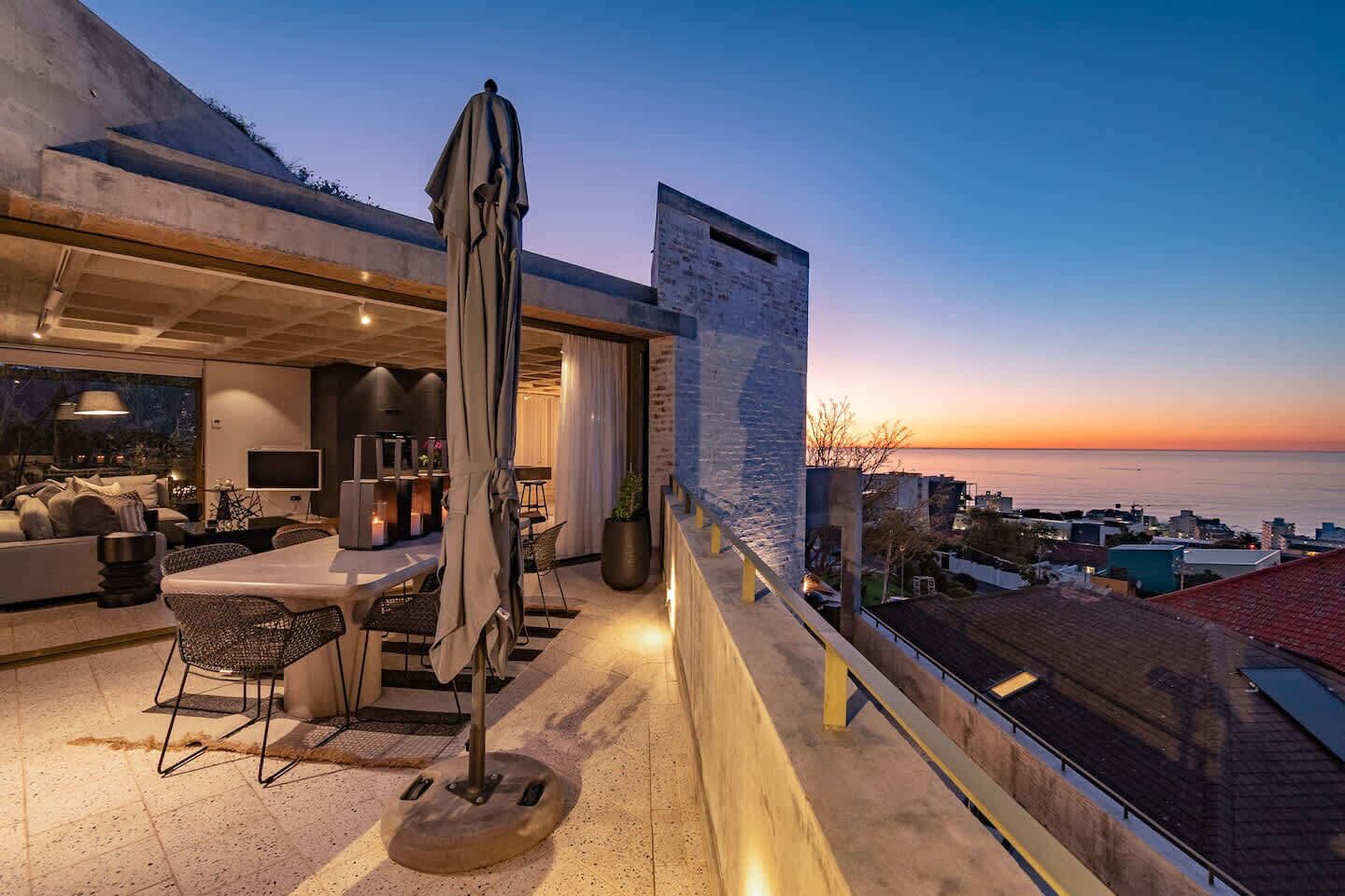Un área de comedor al aire libre con mesa y sillas, un paraguas grande y vista al mar al atardecer.