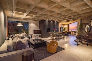 A modern living room with a large sofa, a coffee table, and a TV. There is a kitchen area with a bar and stools. The room has a large window with a view of the sunset. - Luxurious, Pool, Views, Sea Point - Luna Penthouse (Cape Town)