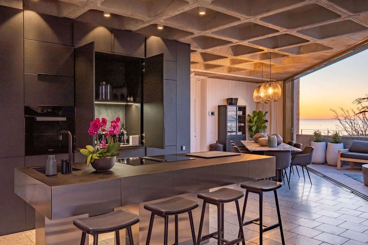 A modern kitchen with a central island, bar stools, and a view of the sea at sunset.