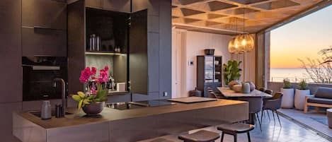 A modern kitchen with a central island, bar stools, and a view of the sea at sunset.