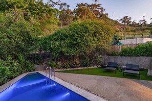 Ein Swimmingpool mit einer Metallleiter, umgeben von einer Betonplattform. Zwei Liegestühle und ein kleiner Tisch sind vorhanden. Das Areal ist von einem Holenzaun und dichter Bepflanzung begrenzt.