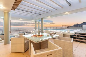 A sunlit outdoor dining area with wicker furniture, a glass table, and a view of the ocean at sunset. - Sunset Azure Penthouse - Panoramic Views, PVT Pool (Cape Town)