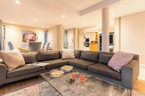 A modern living room with a sectional sofa, a glass coffee table, and a dining area with a kitchen in the background. - Sunset Azure Penthouse - Panoramic Views, PVT Pool (Cape Town)