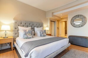 A bedroom with a large bed, two bedside tables with lamps, a bench, and a round mirror on the wall. - Sunset Azure Penthouse - Panoramic Views, PVT Pool (Cape Town)