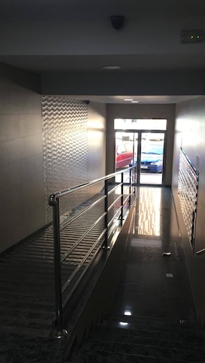 Een gang met een metalen leuning, een structuurwand en zicht op geparkeerde auto's door een glazen deur.