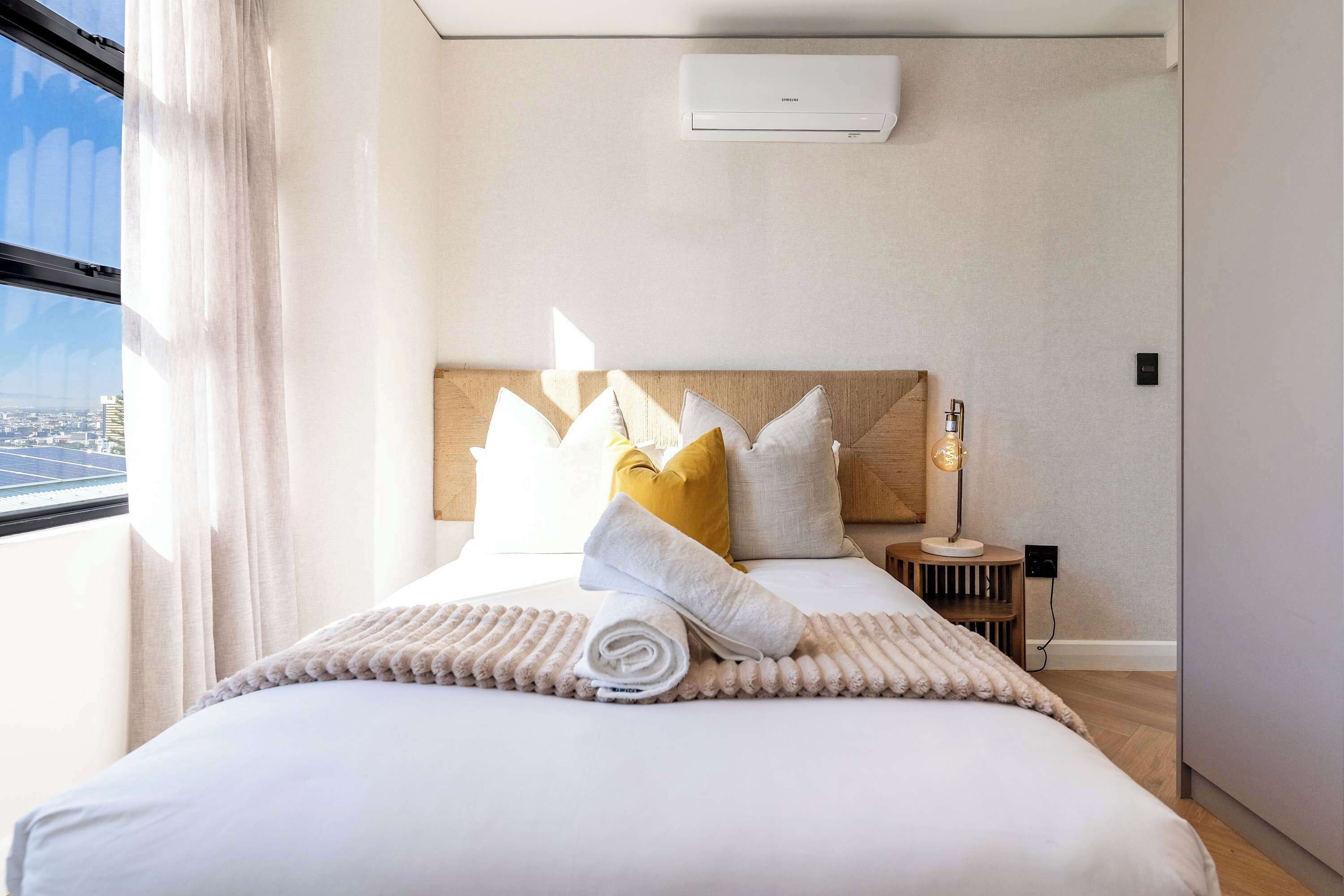 Una habitación de hotel con cama, almohadas, mesita de noche, lámpara y ventana con cortinas.