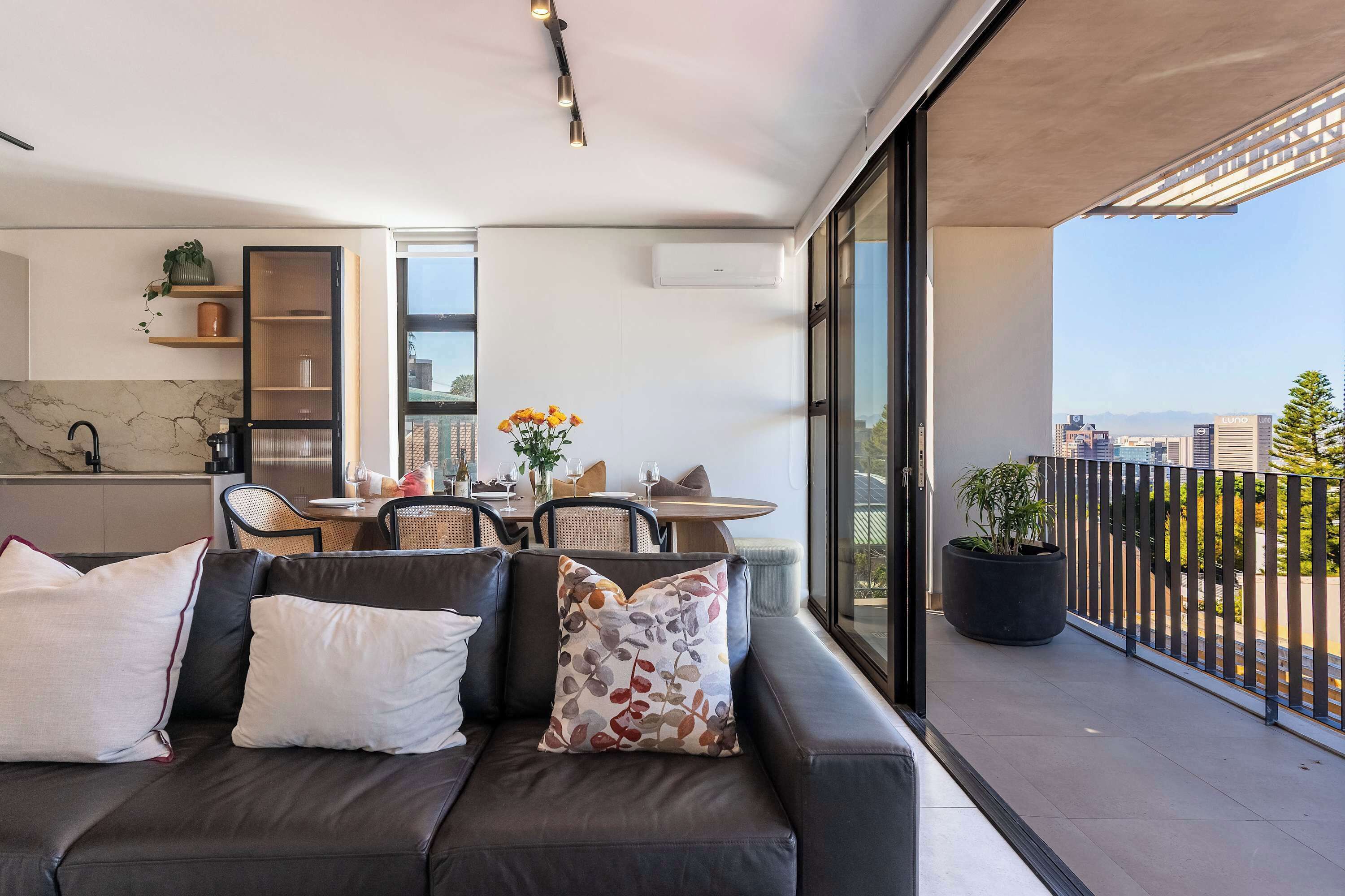 A modern living area with a sofa, a dining table with chairs, a kitchen area, and a balcony with a view of the city.