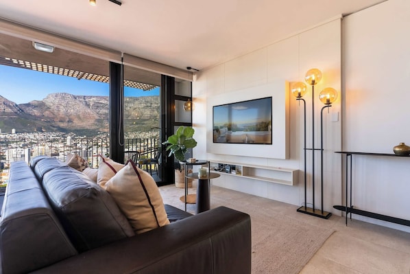 Un salon moderne avec un grand canapé, un téléviseur à écran plat fixé au mur et une vue sur le paysage urbain et les montagnes, visible à travers de grandes fenêtres.