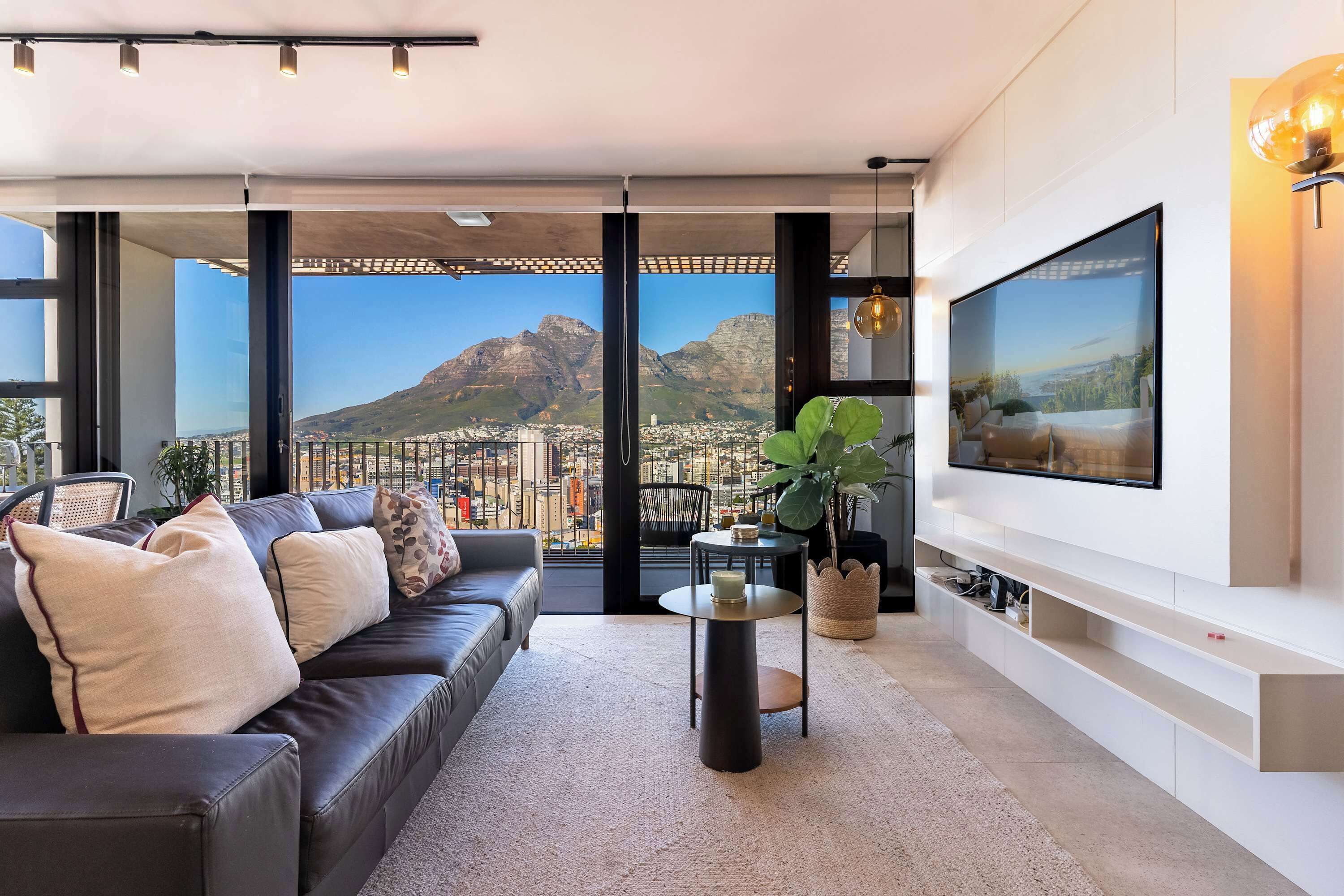 Ein modernes Wohnzimmer mit großem Fernseher, einer Couch, einem Couchtisch und Blick auf eine Stadtlandschaft durch große Fenster.