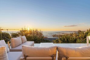 Eine Terrasse mit einer Couch und Stühlen mit Blick auf die Küste bei Sonnenuntergang.