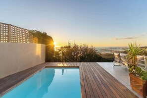 Una piscina con muelle de madera, valla blanca y vista al mar al atardecer.