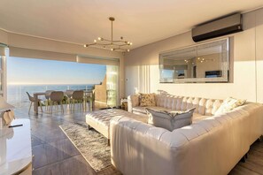 Ein modernes Wohnzimmer mit einem großen weißen Ledersofa, einer Essecke mit Tisch und Stühlen, einem großen Spiegel und Blick aufs Meer.
