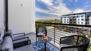 Een balkon met een bank, twee stoelen, een klein rond tafeltje met drank en eten, en uitzicht op de stad.