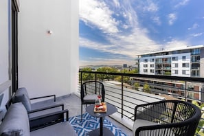 A balcony with a sofa, two chairs, a small round table with a drink and food, and a city view. - Modern & Artistic, Green Point - Urban Palette Apt (Cape Town)