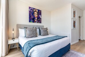 A bedroom with a bed, a nightstand, a lamp, and a painting on the wall. - Modern & Artistic, Green Point - Urban Palette Apt (Cape Town)