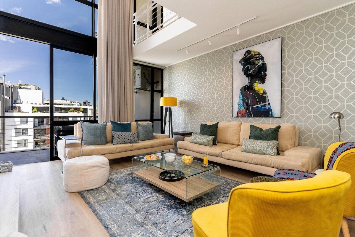 Un salon moderne avec une table basse en verre, deux canapés, un fauteuil jaune et une grande fenêtre donnant sur un paysage urbain.