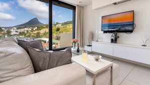 A modern living room with a large window, a flat-screen TV, and a sofa. - Views, De Waterkant CPT, Communal Pool -901 Mirage (Cape Town)