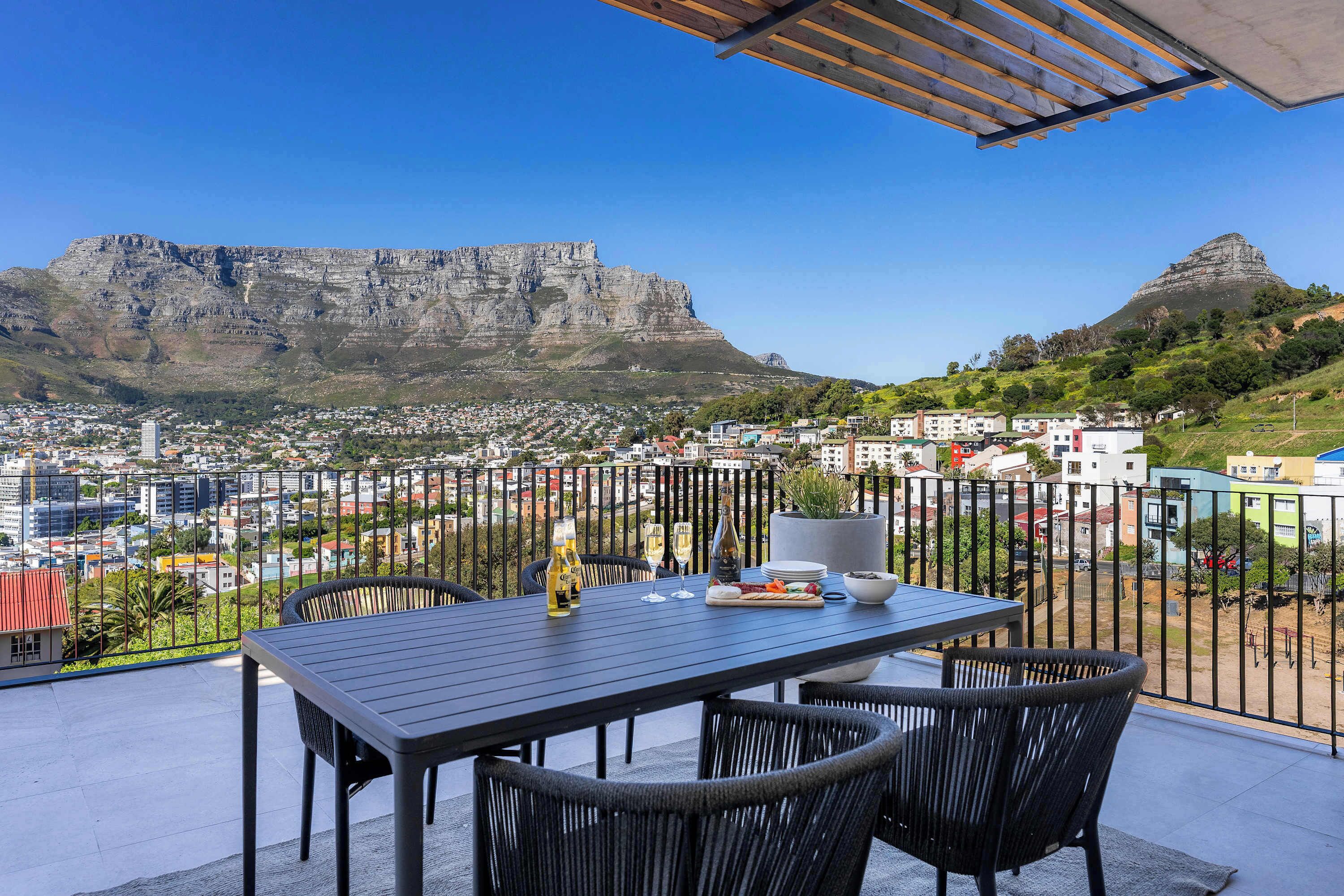 A balcony with a table set for dining, overlooking a cityscape with a prominent mountain in the background.