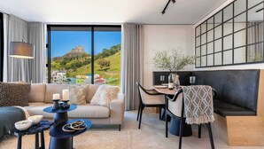 Una sala moderna con sofá, mesa de comedor y sillas, ventanas amplias con vistas a una colina y edificios, y una obra de arte con marco negro en la pared.