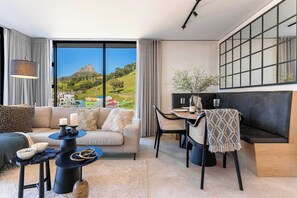 A modern living room with a sofa, dining table, and chairs, large windows with a view of a hill and buildings, and a black-framed artwork on the wall. - Modern, Gorgeous Views, Bo-Kaap - Vista Luxe Apt (Cape Town)