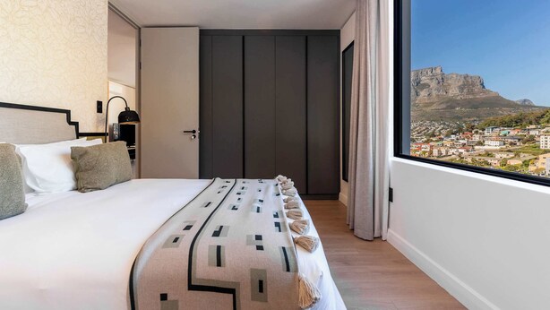 Un dormitorio con un ventanal grande, una cama con cubrecama estampado y vista a un paisaje urbano y montañas.