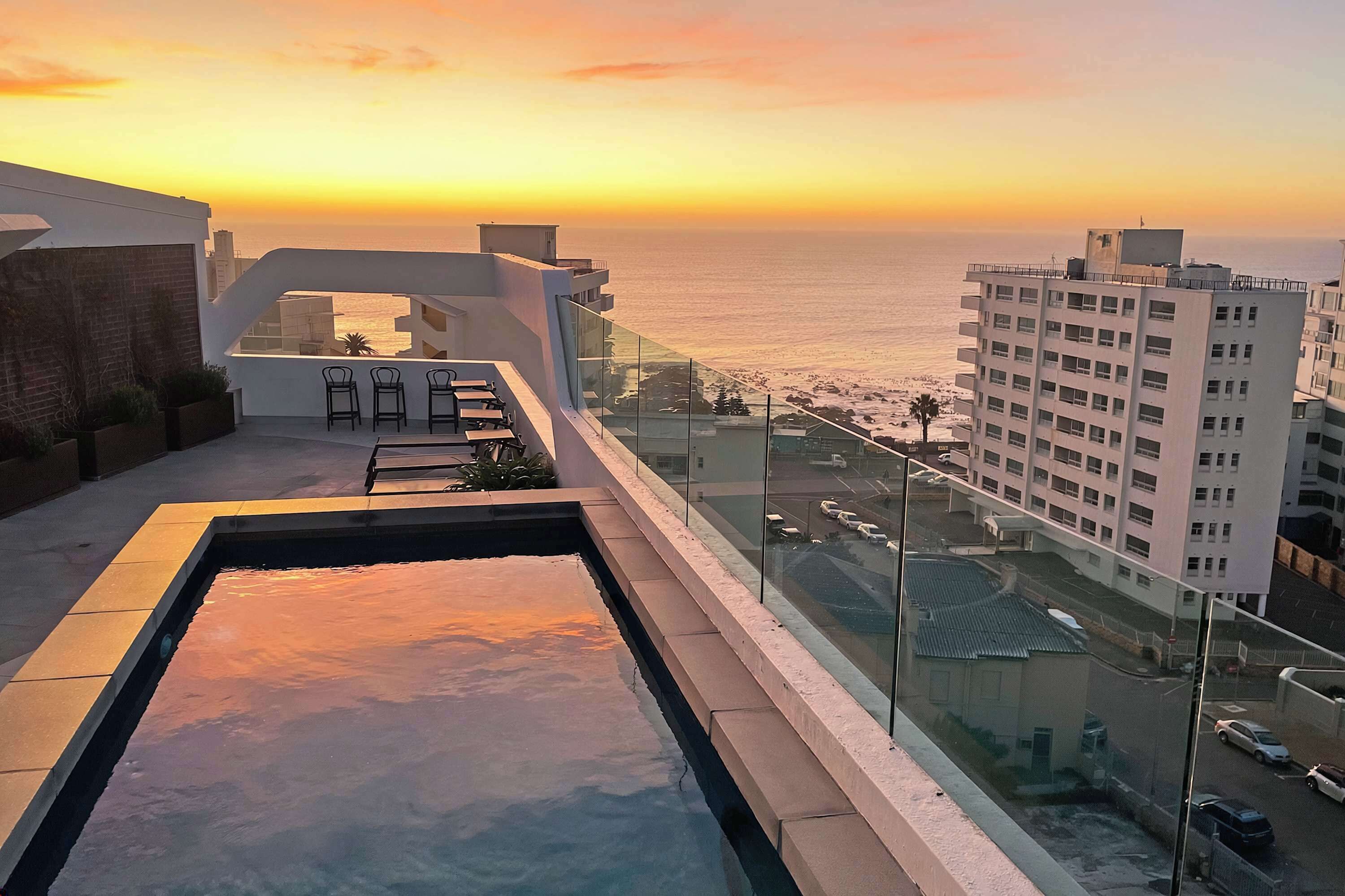 A rooftop pool with a view of the sunset over the ocean, adjacent buildings, and a clear sky.