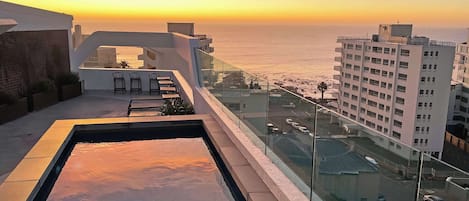 Una piscina en la azotea con vistas a la puesta de sol sobre el océano, edificios cercanos y un cielo despejado.
