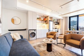 Ein modernes Wohnzimmer mit Sofa, Sesseln, Couchtisch, einer Waschmaschine und einer Küchenzeile mit Regalen und einem Esstisch.