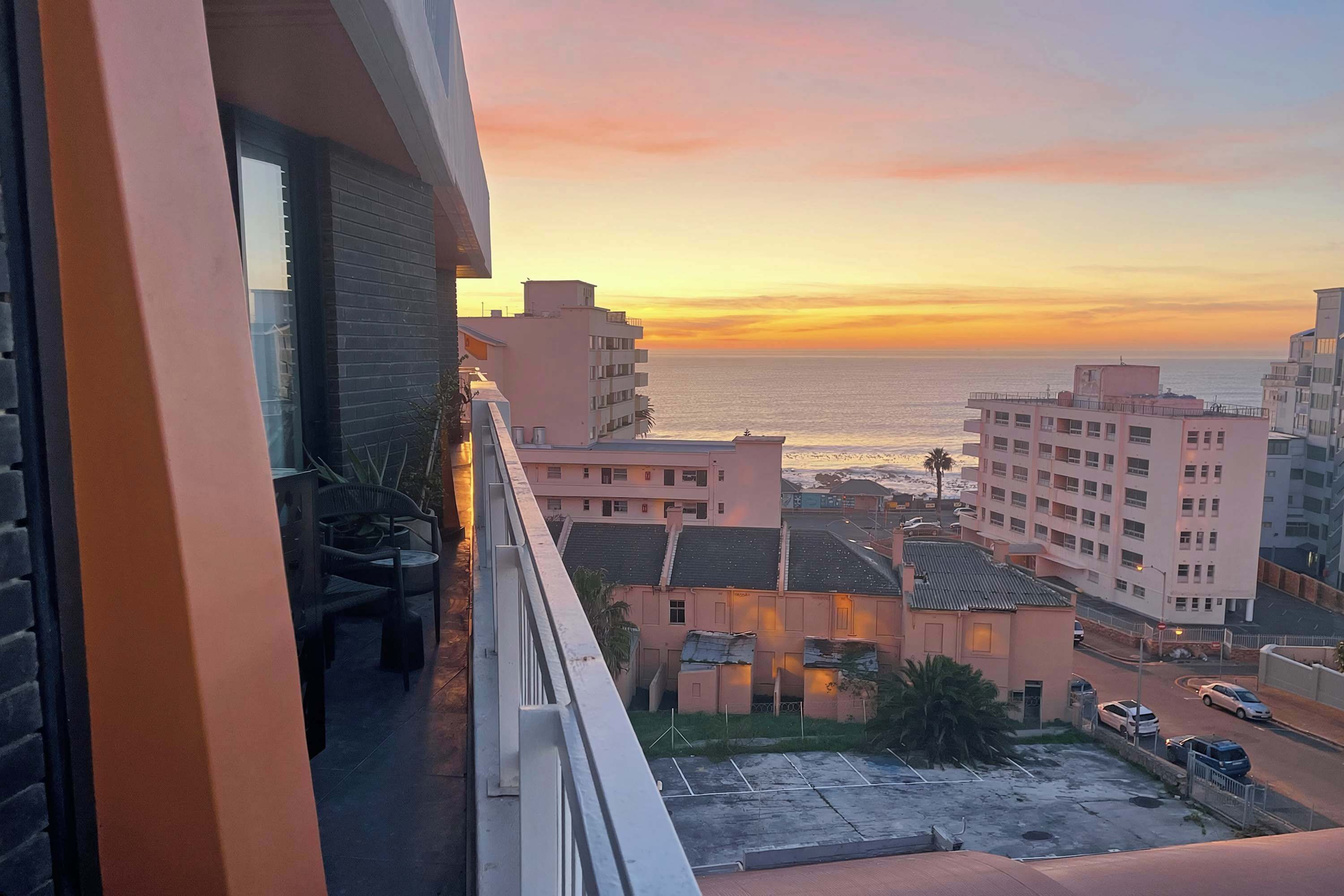 Un balcón con vista al mar al atardecer, edificios en el fondo y un estacionamiento.