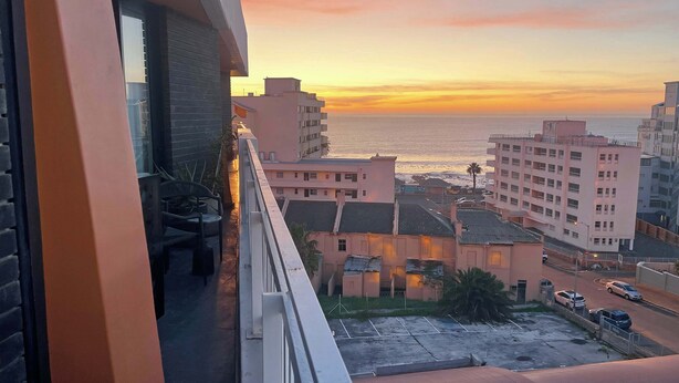 Ein Balkon mit Blick auf das Meer bei Sonnenuntergang, im Hintergrund Gebäude und ein Parkplatz.