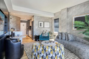 Ein modernes Wohnzimmer mit einer Couch, einem gestreiften Hocker und einem Couchtisch. Im Hintergrund ist eine Küche mit einem Kühlschrank und einer Arbeitsplatte zu sehen.