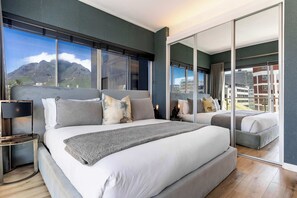 Ein modernes Schlafzimmer mit einem großen Bett, einem Nachttisch, einem Spiegel und Blick auf Berge durch große Fenster.