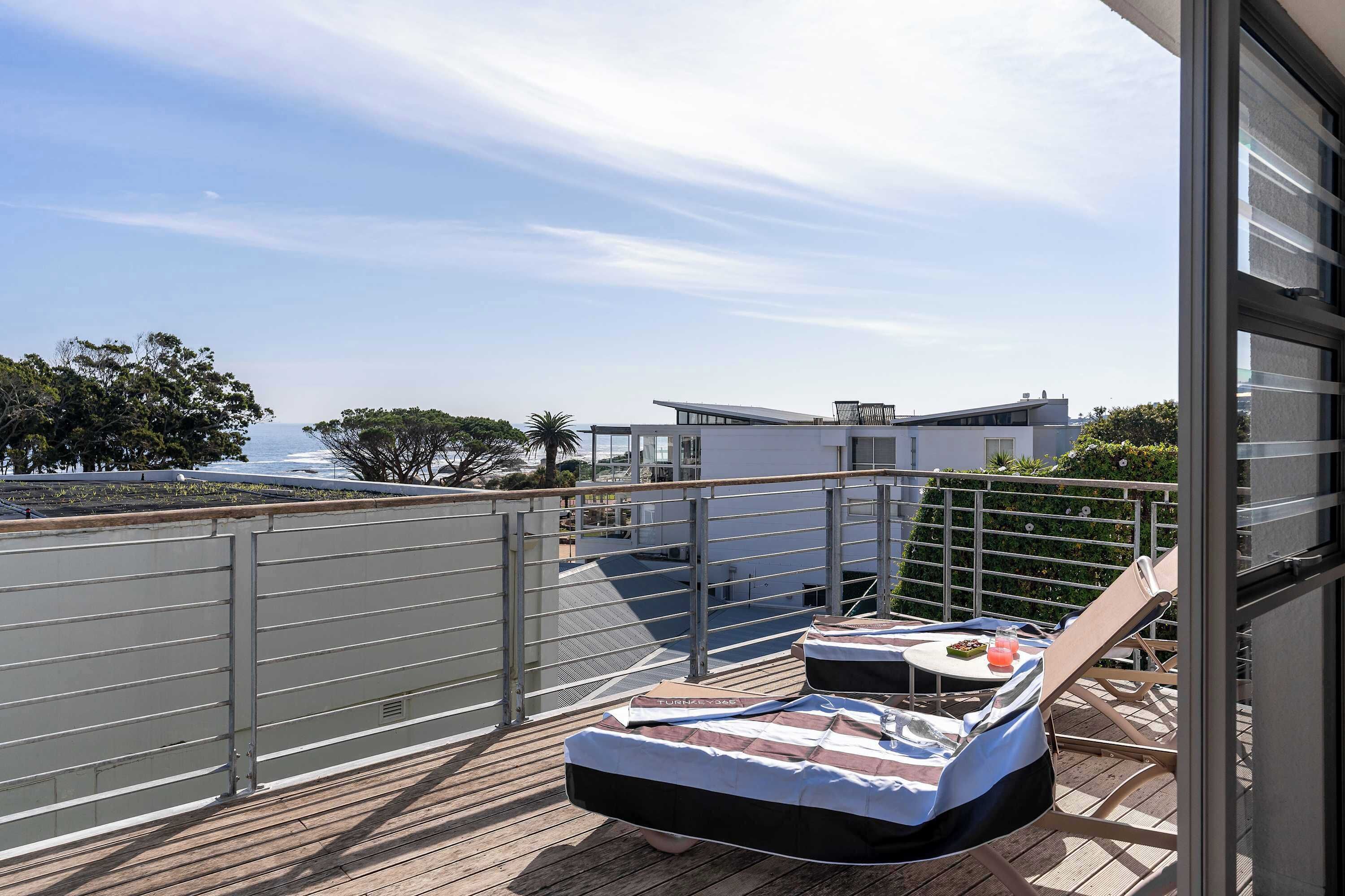 A balcony with a view of the sea, a lounge chair, and a table with a cushion and a glass.