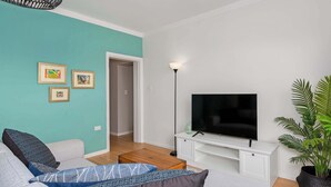 En moderne stue med en teal farvet væg, et fladskærms-tv på et hvidt skab, en sofa med mønstrede hynder og en potteplante.