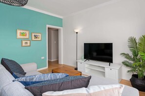 Ein modernes Wohnzimmer mit einer türkisfarbenen Akzentwand, einem Flachbildfernseher auf einem weißen Schrank, einer Couch mit gemusterten Kissen und einer Topfpflanze.