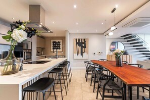A modern kitchen with a central island, bar stools, and a dining area with a wooden table and chairs. - Sea Views, Luxurious, Pool -Iconic View Green Point (Cape Town)