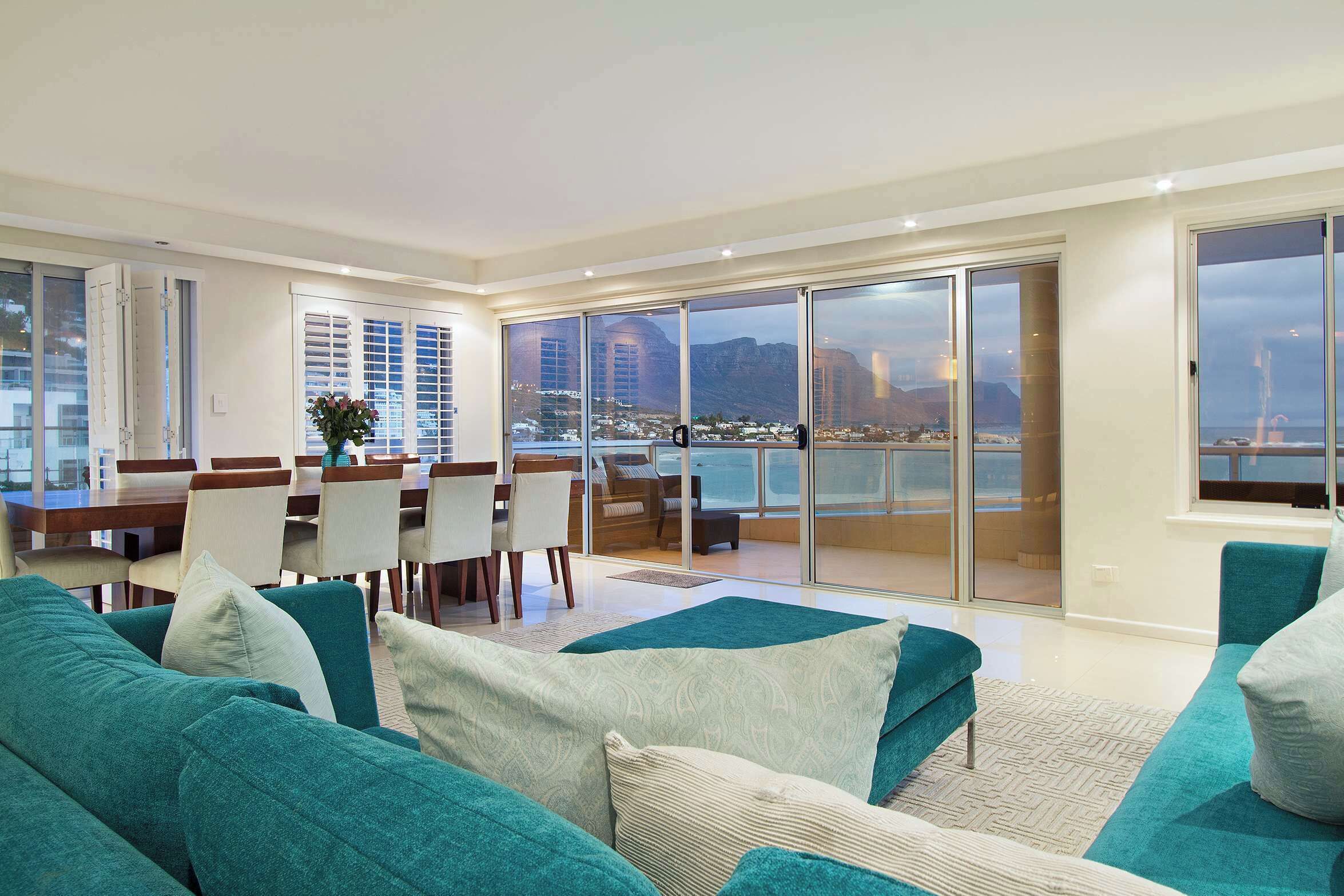 Un salon moderne avec des canapés de couleur turquoise, un coin repas doté d’une table et de chaises en bois, et de grandes portes-fenêtres coulissantes donnant sur un balcon avec vue sur la ville.