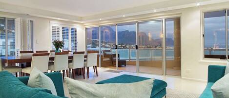 A modern living room with teal sofas, a dining area with a wooden table and chairs, and large sliding glass doors leading to a balcony with a city view.