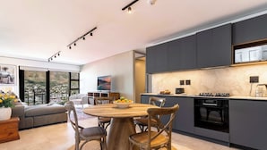 A modern kitchen with dark cabinets, a wooden dining table, and a large window overlooking a scenic view. - 270 Views, Modern, Bo-Kaap -Table Mountain Majesty Apt (Cape Town)